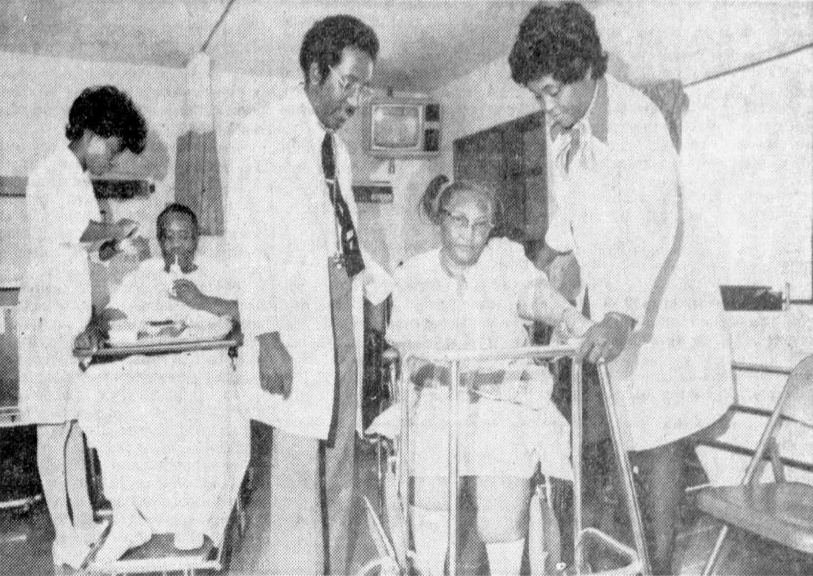 A doctor and two nurses stand over two seated patients in a hospital room with a small television mounted in the corner of the room